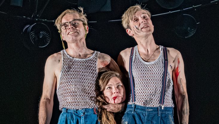 Büstenansicht von zwei Männern auf einer Bühne, der linke hält einen Frauenkopf unter dem Arm